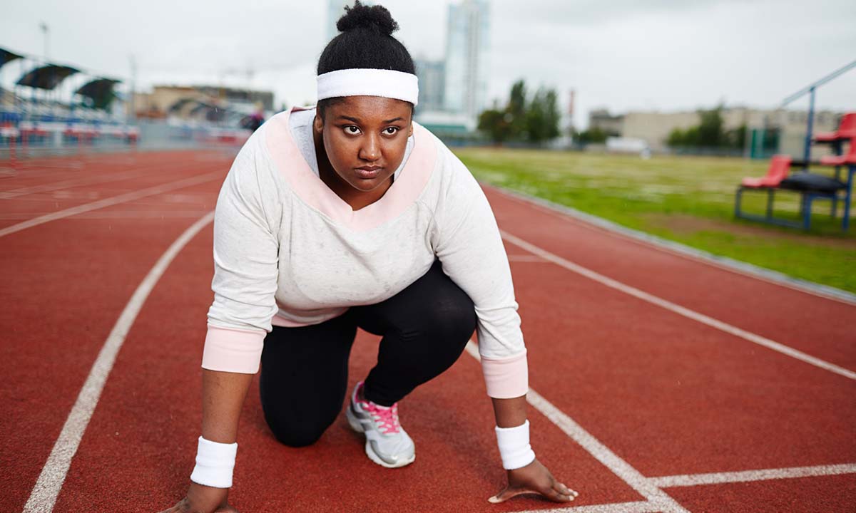 A young black woman is knelt at the start line of an outdoor race track