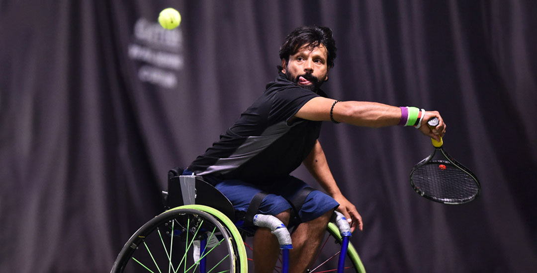 A middle ahed man with dark hair and a beard takes a swing at a tennis ball from his wheelchair. 